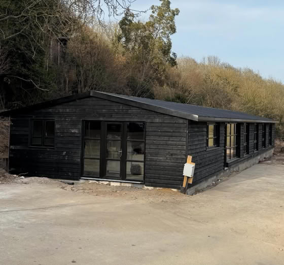 A long, dark wooden building with multiple windows, situated on a dirt surface with trees in the background.