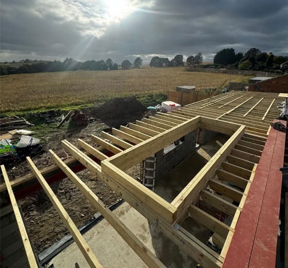 Wooden beams forming the framework of a roof under construction, with a ladder and tools visible, set against a backdrop of a field and cloudy sky with the sun shining through.
