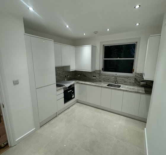 A modern kitchen with white cabinets, a built-in oven, a stove, and a sink under a window. The floor is tiled, and the ceiling has recessed lighting.