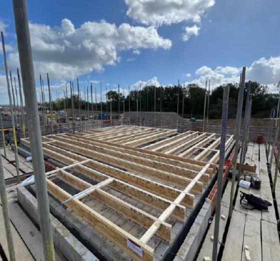 Construction site with wooden framework on a concrete foundation, surrounded by scaffolding poles under a partly cloudy sky.