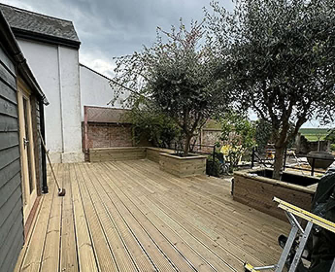 Wooden deck with trees and plants in planters, adjacent to a building with dark siding and a white wall, under a cloudy sky.