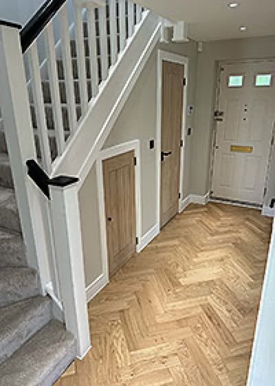 A hallway with a staircase on the left, wooden flooring in a herringbone pattern, and three closed interior doors, including a new front door.