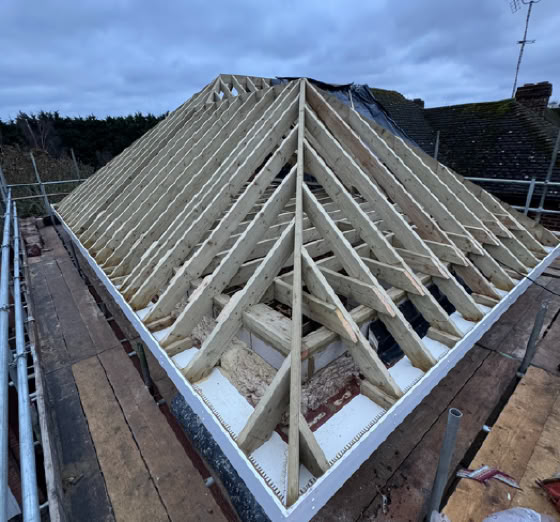 Wooden roof trusses under construction on a building, with scaffolding around the perimeter and a cloudy sky above.