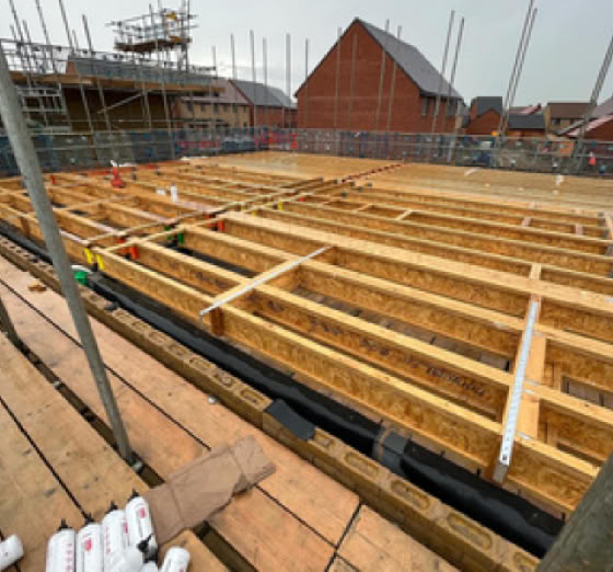 Construction site with wooden framework on the ground, surrounded by scaffolding and partially built structures in the background.