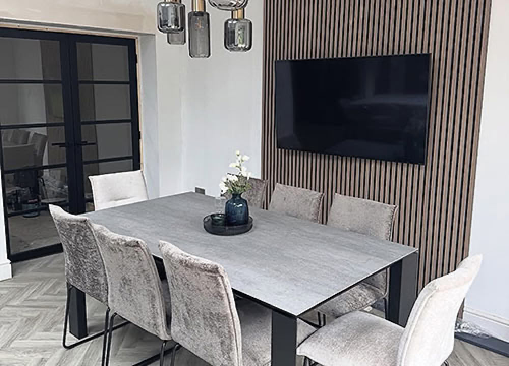 A modern dining room renovation with a rectangular table surrounded by six upholstered chairs, a wall-mounted TV, decorative wooden panelling, and pendant lights hanging above.