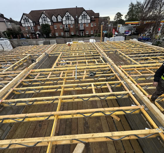 Construction site with wooden framework, a person in a high-visibility vest, and residential buildings in the background.