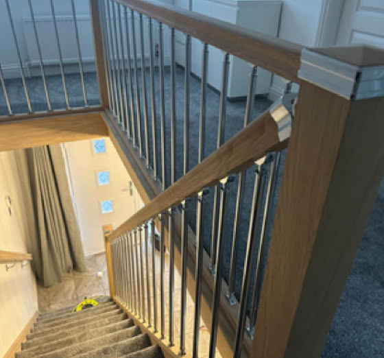 Carpeted staircase with wooden handrails and metal balusters, leading down to a lower floor with a window and a door visible in the background.