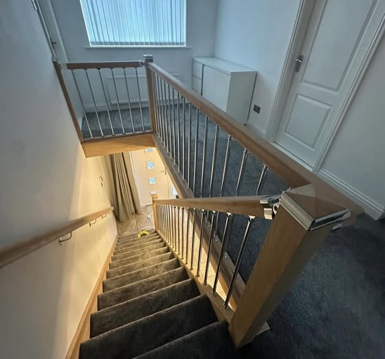 A staircase with wooden handrails and carpeted steps, leading down to a lower floor, with a window and a closed door visible at the top landing.