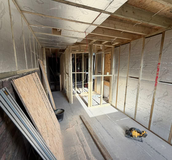 A partially constructed interior space with exposed wooden framing, insulation on the walls, and construction materials on the floor.