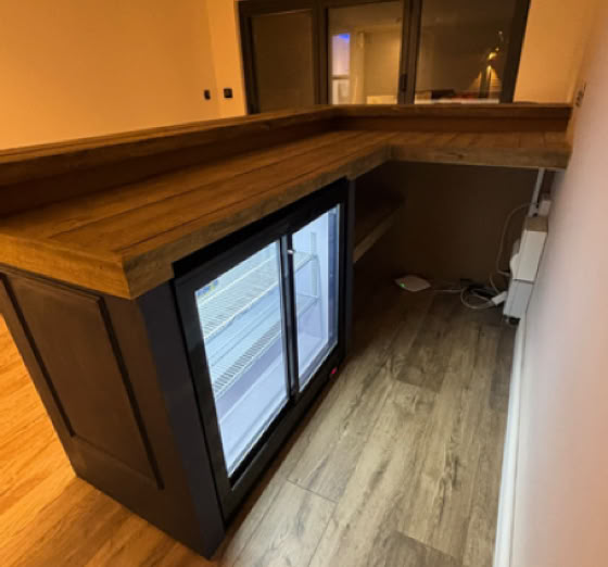 Wooden bar counter with a glass-front refrigerator underneath, set in a room with wooden flooring and a window in the background.