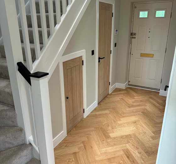 A hallway with light wood herringbone flooring, a staircase with carpeted steps and white railings, a small door under the stairs, and a main entrance door with a mail slot and two small windows.