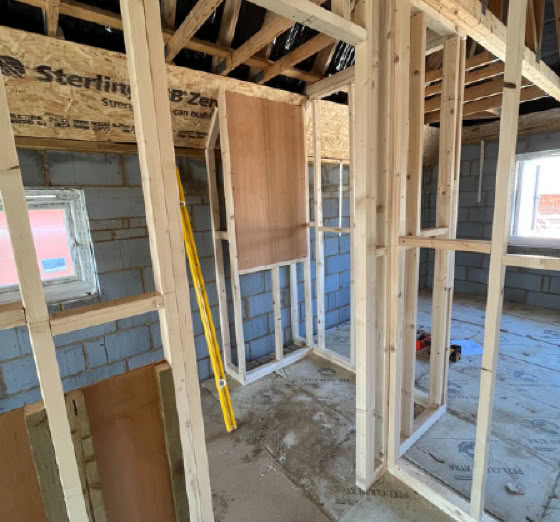 Interior of a building under construction with exposed wooden framing, unfinished walls, and a yellow ladder leaning against a wall.