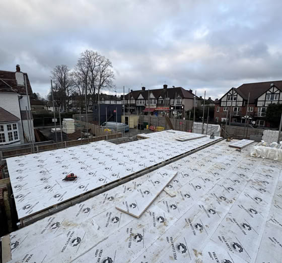A construction site with a flat roof covered in insulation panels, surrounded by residential houses and trees under a cloudy sky.